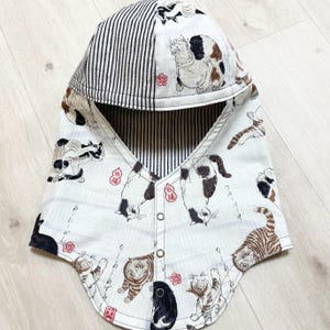 May include: A hooded neck warmer featuring a white fabric base with a repeating pattern of various cats in brown, black, and white. The hood interior and trim feature black and white vertical stripes. The front includes three silver metal snap buttons.