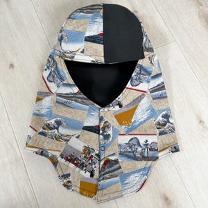 May include: A balaclava-style head covering with a black top and a patterned front. The pattern features a collage of Japanese art with waves, mountains, and figures in shades of blue, beige, and brown. The head covering has a button closure.