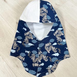 Può includere: Un passamontagna blu navy con cappuccio bianco. Il passamontagna presenta un motivo ripetuto di draghi e nuvole bianche. Il passamontagna ha una chiusura con bottoni sul collo ed è realizzato in un tessuto morbido.