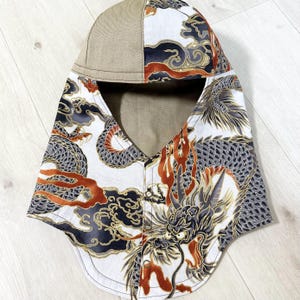 May include: A hooded head covering featuring a tan solid-colored top panel and a white base fabric printed with a detailed dragon motif in shades of black, gray, and burnt orange. The design includes stylized clouds and a metal snap closure.