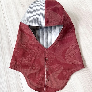 May include: A hood-style head covering featuring a deep red fabric with a subtle geometric pattern on the exterior and a navy blue and white pinstriped lining. The front closure includes three small circular buttons aligned vertically along the center.