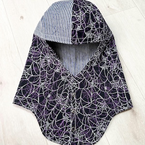May include: A hooded neck warmer featuring a black fabric with a white and purple spiderweb pattern. The hood has a contrasting section of blue and white vertical stripes. The front closure includes three silver-toned metal snaps for fastening.
