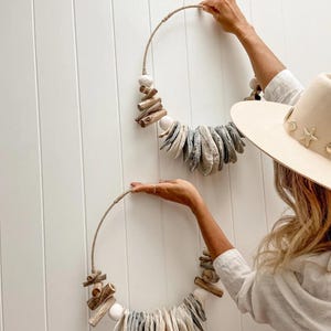 May include: Two circular wall hangings crafted from natural materials. Each features a rope frame decorated with driftwood, white stones, and grey-toned shell pieces. The hangings are displayed against a white, slatted wall, reflecting a coastal or bohemian style.