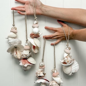 May include: Four hanging ornaments crafted from natural seashells and sea urchin tests in shades of white, cream, and soft pink. Each ornament is strung on a piece of tan jute twine, featuring a variety of textured shell shapes and sizes.