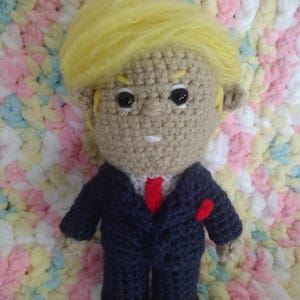 May include: A handmade crochet doll of a man wearing a navy blue suit with a red tie and a red pocket square. The doll has a tan face, black eyes, and yellow hair. The doll is on a pastel-colored blanket.