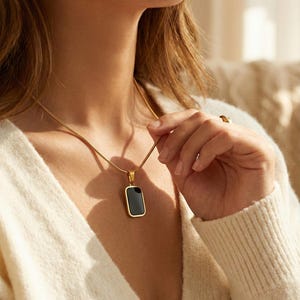 May include: Gold necklace featuring a rectangular black pendant. The pendant has a gold border and hangs from a fine chain. The necklace is worn over a cream-coloured jumper.