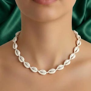 Puede incluir: Un collar gargantilla hecho de conchas blancas. Las conchas ovaladas están ensartadas, creando un aspecto natural y playero. El collar se muestra sobre un fondo de tela verde. Las conchas miden aproximadamente 2-3 cm de largo.