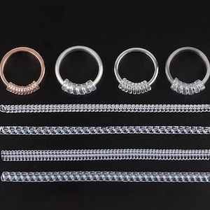 Puede incluir: Una colección de anillos de plata y oro rosa con tallas de anillos de plástico transparente. Los anillos tienen un diseño de banda simple con una serie de pequeñas cuentas de plástico transparente. También se muestran varias tiras de tallas de anillos de plástico transparente.