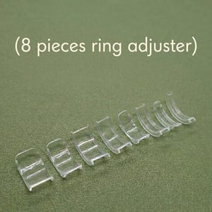 Puede incluir: Ocho ajustadores de anillos de plástico transparente están dispuestos sobre una superficie verde. Los ajustadores son curvos y tienen un diseño similar a una escalera. El texto superior dice "(8 pieces ring adjuster)".