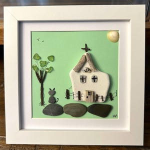 May include: A framed pebble art piece featuring a white church with a cross, a tree with green leaves, and a gray cat. The background is light green, and the frame is white. A yellow shell represents the sun.