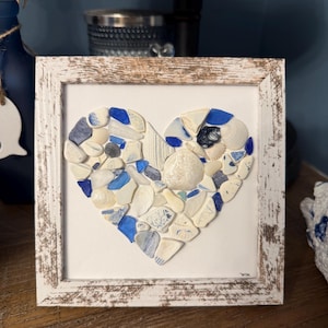 May include: A square wall art piece featuring a heart-shaped mosaic made of various white and blue sea glass, ceramic shards, and small seashells. The mosaic is set against a white background and enclosed in a distressed, weathered white wooden frame.