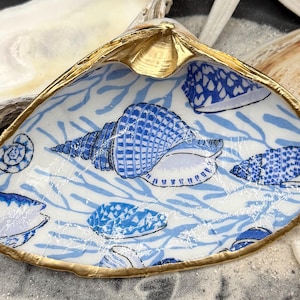 May include: A decorative seashell dish with a gold rim and a blue and white seashell pattern. The dish features a large, detailed seashell design in the center, surrounded by smaller seashell motifs and seaweed-like patterns.