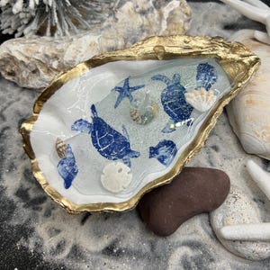 May include: A decorative oyster shell with a gold-painted rim, filled with a clear resin scene featuring blue sea turtles, starfish, and seashells. The shell rests on a bed of sand with additional decorative elements.