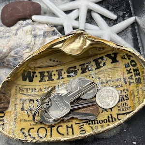 May include: A decorative, gold-edged clam shell dish with the word "WHISKEY" printed on the interior. The dish holds keys and coins. Starfish and a small rock are in the background. The dish has a vintage, rustic aesthetic.