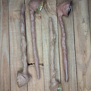 May include: Four handcrafted, rustic-style tobacco pipes. The pipes are primarily brown with textured stems and bowls. Some bowls have green or gold accents. The pipes are arranged on a wooden surface.