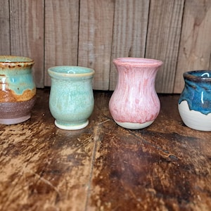 May include: Four handmade ceramic vessels in varying colors. The first is brown with turquoise and brown accents. The second is light green. The third is pink. The fourth is white with blue accents. Each has a unique shape and glaze.