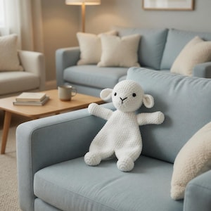 Könnte beinhalten: Ein weißes Häkellamm-Spielzeug mit schwarzen Augen und einer schwarzen 'Y'-förmigen Nase, das auf einem hellblauen Sofa liegt. Das Lamm hat ausgestreckte Arme und kleine, abgerundete Ohren. Der Hintergrund zeigt ein Wohnzimmer.