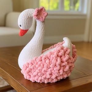 Könnte beinhalten: Ein handgefertigter Schwan aus weißer und rosa Häkelarbeit. Der Schwan hat einen weißen Körper, eine rosa Blume auf dem Kopf und einen roten Schnabel. Der Schwan sitzt auf einem Holztisch.