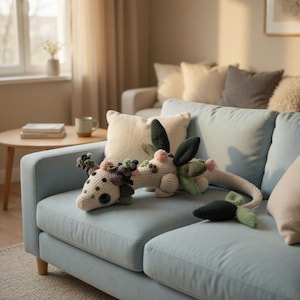 Könnte beinhalten: Zwei gehäkelte Drachenspielzeuge in Creme-, Grün- und Rosatönen liegen auf einem hellblauen Sofa. Die handgefertigten Spielzeuge weisen detaillierte Designs auf, darunter blattartige Akzente und ein langer Schwanz. Die Szene spielt in einem gemütlichen Wohnzimmer.