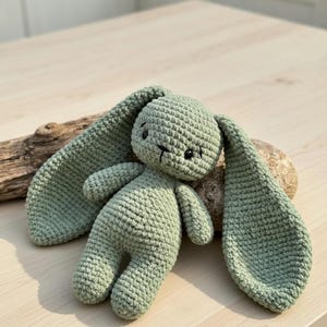 Könnte beinhalten: Ein handgefertigtes, salbeigrünes Häschen aus Häkelarbeit mit langen, schlaffen Ohren. Das Plüschtier hat ein einfaches, süßes Gesicht mit schwarzen Augen und einem gestickten Mund. Das Häschen liegt auf einer hellen Holzoberfläche, neben einem Stück Treibholz und einem kleinen Stein.