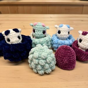 Könnte beinhalten: Eine Sammlung handgefertigter Häkeltiere in verschiedenen Farben. Die Kreaturen haben kleine Hörner und große Augen. Zu den Farben gehören Weiß, Blau, Grün und Lila. Die Kreaturen sind in farblich passenden Nestern oder Hülsen eingebettet.