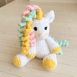 May include: A white crochet unicorn plush toy with a colorful mane in pastel shades of pink, yellow, and green. The unicorn has black eyes and yellow accents on its horn and hooves. The toy is sitting on a light-colored wooden surface.