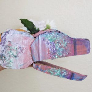 May include: A handmade, whimsical dragon head mask crafted from cardboard, painted in shades of pink, purple, and blue with iridescent accents. The mask is adorned with white flowers and green leaves, creating a fantasy-themed accessory.