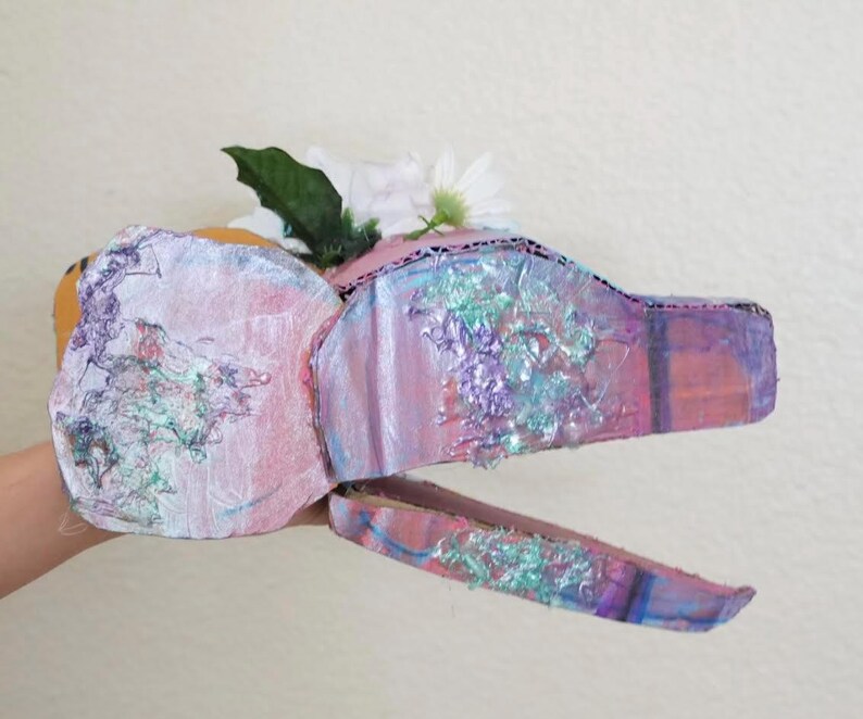 May include: A handmade, whimsical dragon head mask crafted from cardboard, painted in shades of pink, purple, and blue with iridescent accents. The mask is adorned with white flowers and green leaves, creating a fantasy-themed accessory.