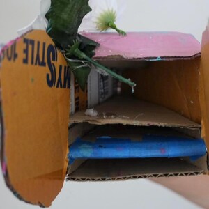 May include: A handmade cardboard box with a pink top and two interior shelves. The box is decorated with artificial flowers and the text "MY STYLE 10". The interior contains a blue object.