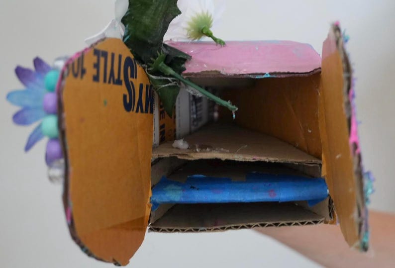 May include: A handmade cardboard box with a pink top and two interior shelves. The box is decorated with artificial flowers and the text "MY STYLE 10". The interior contains a blue object.