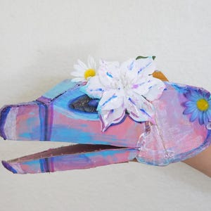 May include: A handmade, colorful dinosaur puppet with a painted cardboard construction. The puppet features shades of pink, blue, and purple, adorned with white and blue artificial flowers. The puppet is held by a person.