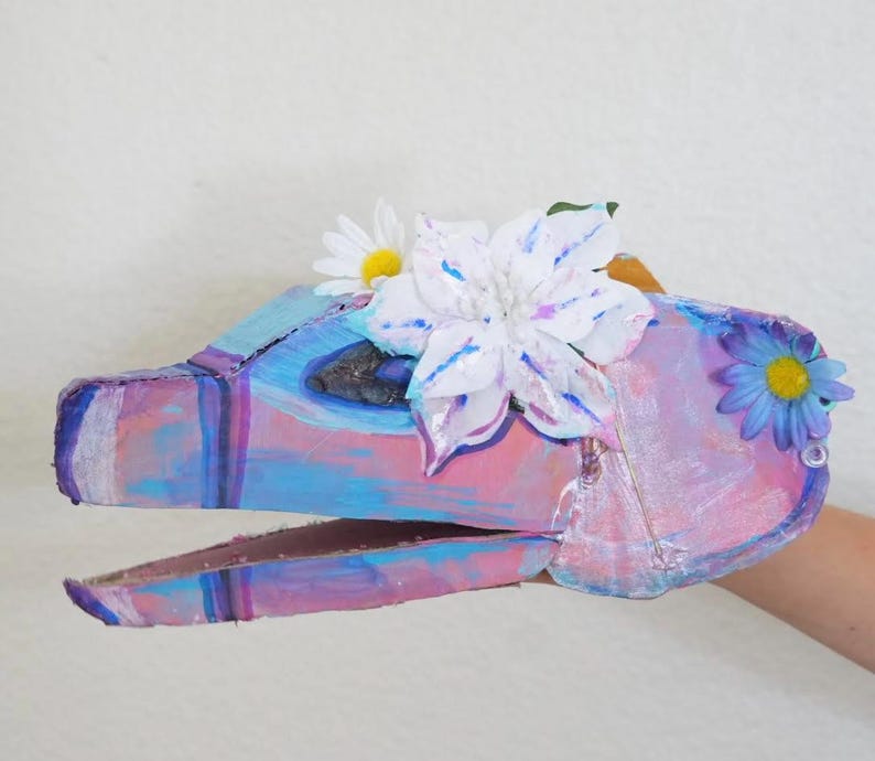 May include: A handmade, colorful dinosaur puppet with a painted cardboard construction. The puppet features shades of pink, blue, and purple, adorned with white and blue artificial flowers. The puppet is held by a person.