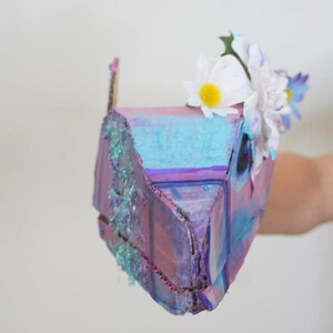 May include: A handmade, colorful cardboard box painted with shades of blue, purple, and pink. The box is decorated with artificial white and blue flowers. The box has a rustic, textured appearance.