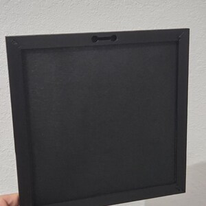 May include: A square black picture frame, held by a hand, with a keyhole slot for hanging. The frame has a matte finish and a simple, modern design. The back of the frame is visible, showing the construction and materials.