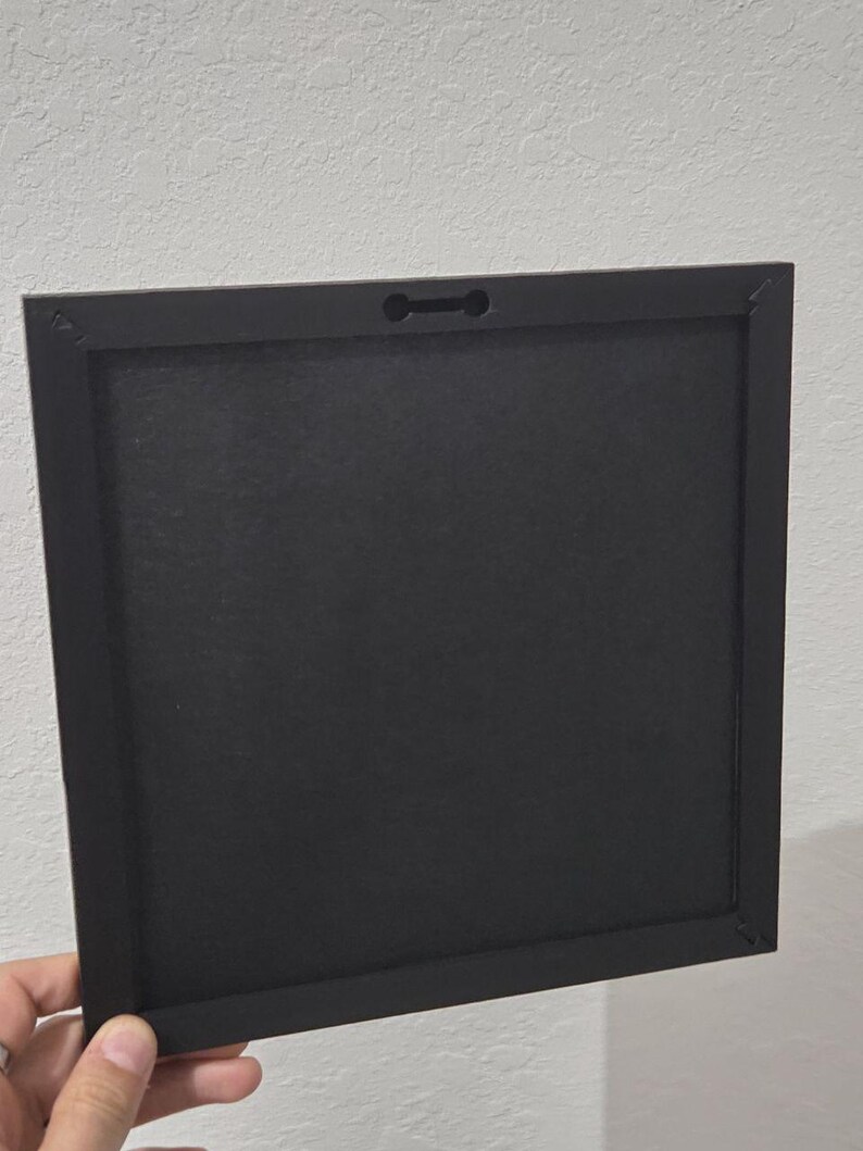 May include: A square black picture frame, held by a hand, with a keyhole slot for hanging. The frame has a matte finish and a simple, modern design. The back of the frame is visible, showing the construction and materials.