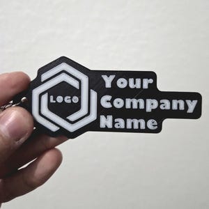 Könnte beinhalten: Schlüsselanhänger in Schwarz und Weiß mit dem Text "Your Company Name" und einem Logo. Der Schlüsselanhänger wird von einer Hand gehalten. Das Logo ist ein weißes Sechseck in einem Sechseck, mit dem Wort "LOGO" darin.