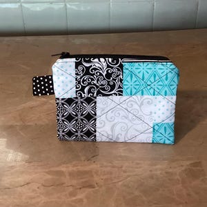 May include: A small, quilted pouch with a black zipper and a black and white polka dot tab. The pouch is made of fabric squares in black, white, and teal, featuring various patterns. The pouch is approximately 6 inches wide and 4 inches tall.