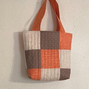 May include: A quilted tote bag with orange handles and a patchwork design. The bag features squares of beige, brown, and orange fabric, some with text. The bag is hanging against a white wall.