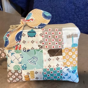 May include: A patchwork zippered pouch with a coordinating bow. The pouch features a variety of patterned squares in shades of blue, yellow, brown, and white. A silver-colored clasp is attached to the side.