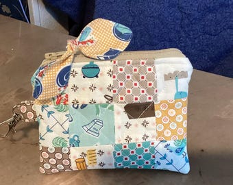 May include: A patchwork zippered pouch with a coordinating bow. The pouch features a variety of patterned squares in shades of blue, yellow, brown, and white. A silver-colored clasp is attached to the side.