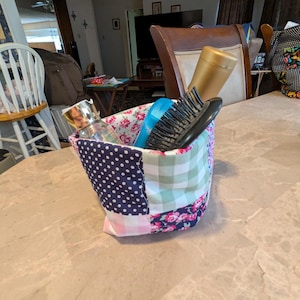 May include: A patchwork fabric basket with a variety of patterns, including polka dots, gingham, and floral prints. The basket contains a hairbrush, a bottle of shampoo, and other toiletries. The basket is sitting on a marble countertop.