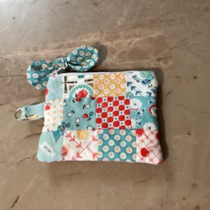 Puede incluir: Un monedero de patchwork con un tirador de cremallera en forma de lazo a juego. El bolso presenta una variedad de cuadrados estampados en tonos de azul, rojo, amarillo y blanco. Tiene un anillo de metal plateado adjunto en el lateral.