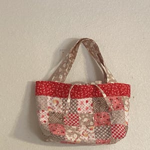 May include: A quilted tote bag with a patchwork design. The bag features a red top border and handles, with a body composed of various squares in shades of red, pink, and beige. The bag is designed for carrying items.