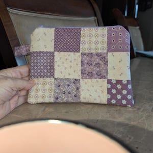 May include: A patchwork zippered pouch in shades of purple and cream. The pouch is made of small fabric squares with floral and geometric patterns. A small fabric loop is attached to the side. The pouch is held by a hand.