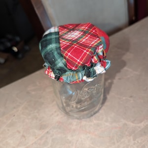 May include: A clear glass jar topped with a fabric cover featuring a patchwork design of red, green, and white plaid patterns. The fabric is gathered with an elastic band around the rim of the jar, creating a ruffled edge that sits securely.