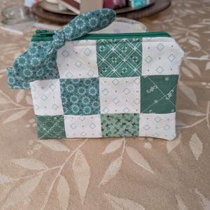 Puede incluir: Un pequeño monedero patchwork hecho a mano con un tirador de cremallera de tela a juego. El monedero presenta un patrón de cuadrados blancos y verdes con varios diseños geométricos. El tirador de la cremallera está anudado. El monedero está sobre un mantel beige.