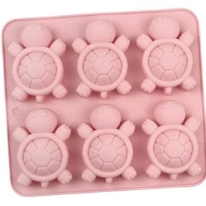 May include: A rectangular light pink silicone mold featuring six identical cavities shaped like turtles. Each turtle cavity displays a rounded head, four small limbs, and a shell with a distinct hexagonal pattern embossed on the surface.