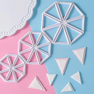 May include: Three white plastic circular cookie cutters with internal dividers that create eight triangular segments, accompanied by several small white triangular plastic pieces, arranged on a split pink and light blue background.
