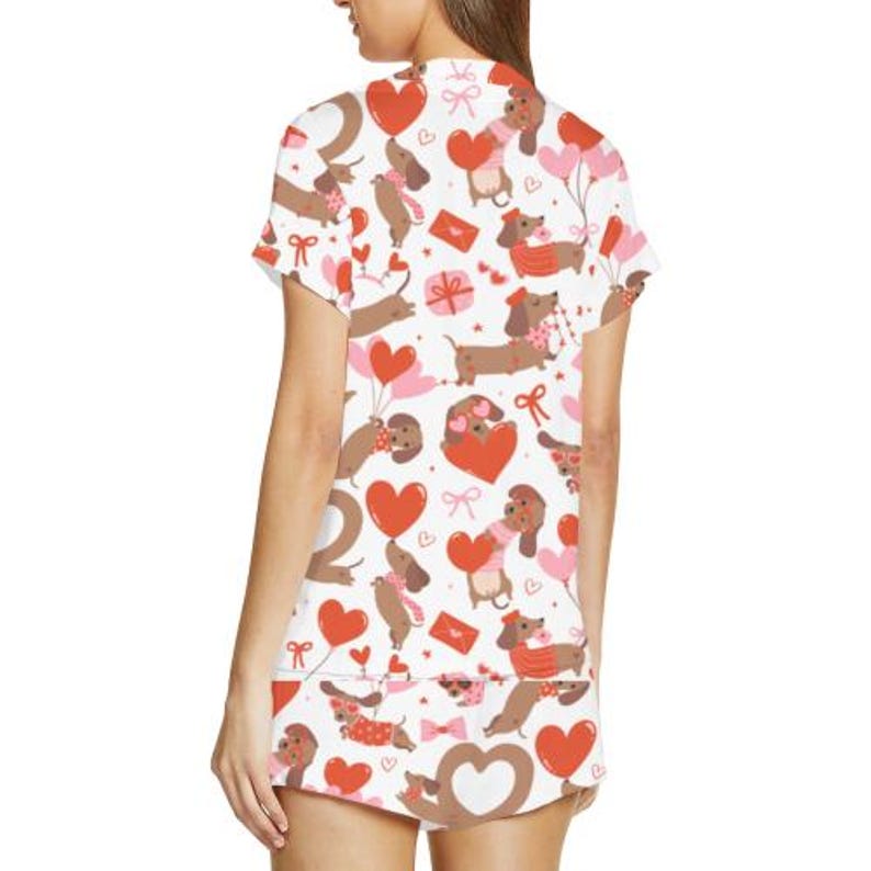 May include: White pyjama set with a repeating pattern of brown dachshunds, red hearts, pink balloons, and bows. The short-sleeved top and shorts feature a Valentine's Day theme.