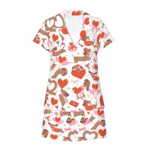 May include: A white, short-sleeved dress with a repeating pattern of brown dachshunds, red hearts, and pink balloons. The dress has a V-neck collar and button closure. The design is festive and playful, suitable for a special occasion.